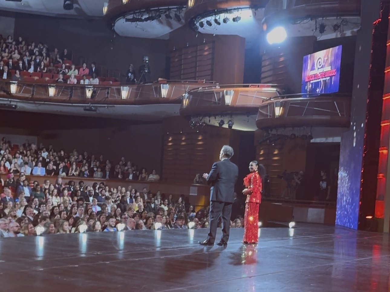 Chelsea and her co-host on stage in front of a packed theater during the live awards broadcast
